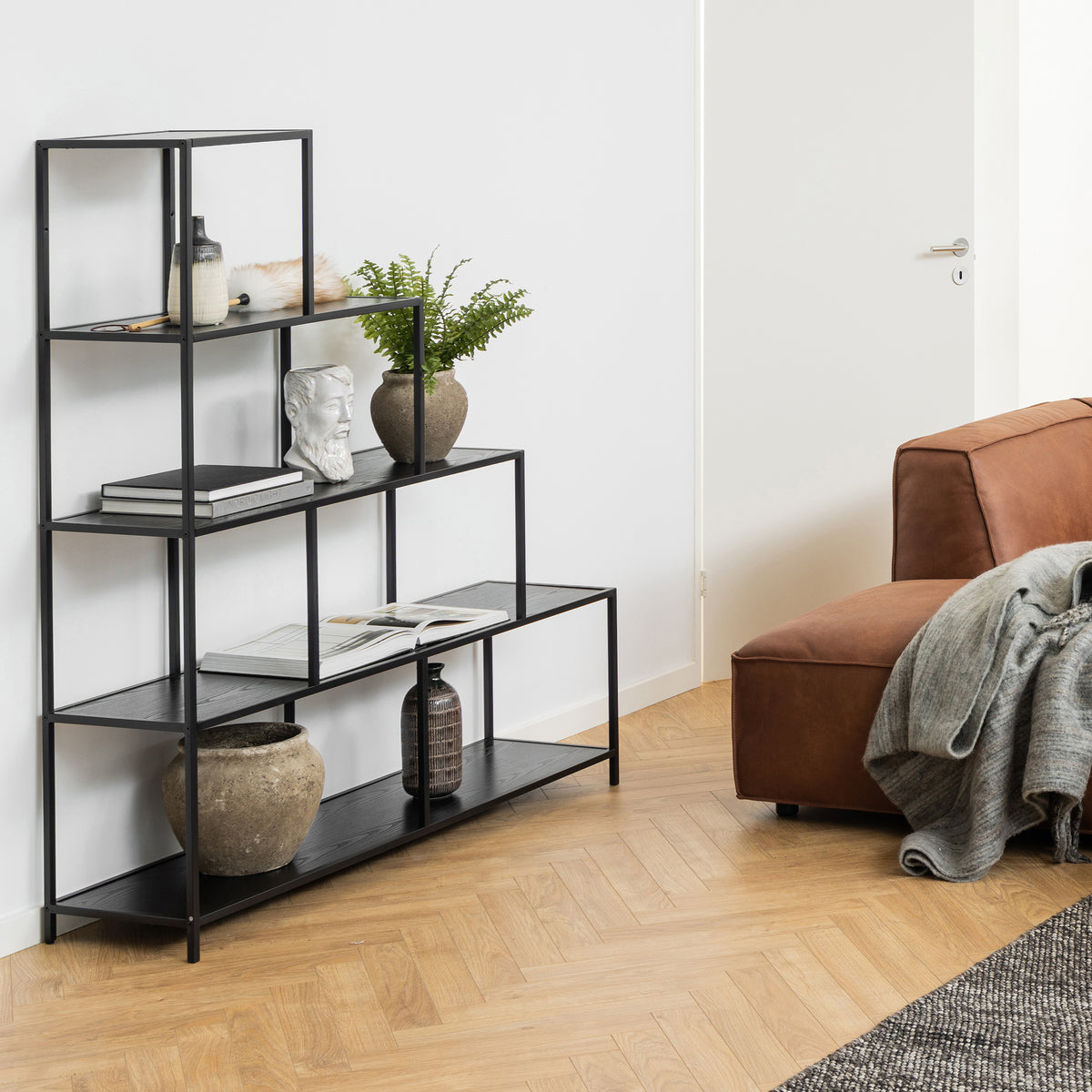 The Skeleto bookcase (135x150), in black melamine, in the living room next to a brown sofa, with a plant, a bust and books