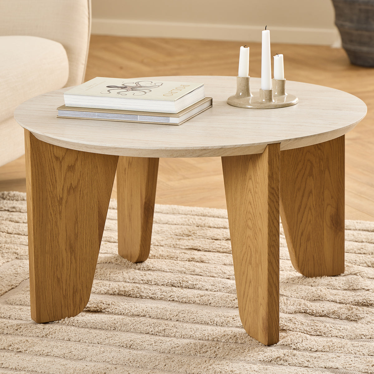 The Tendra coffee table (Ø80x45) on a beige rug next to a light-coloured armchair, with books and a candlestick – a coffee table for the living room