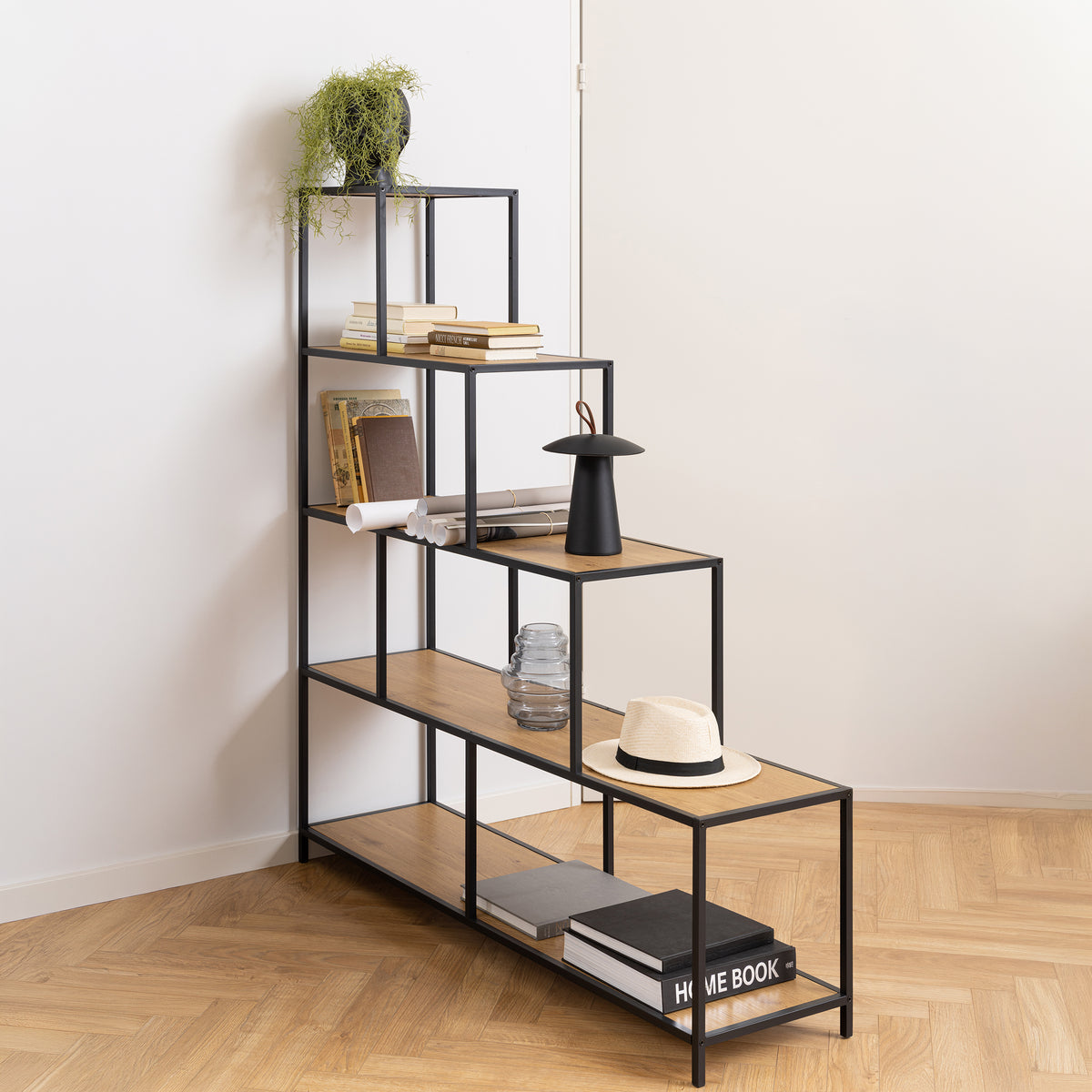 Skeleto bookcase, 135x150, Wild Oak, in the corner of the room on a herringbone floor, with books, a lamp and a hat – a stepped bookcase