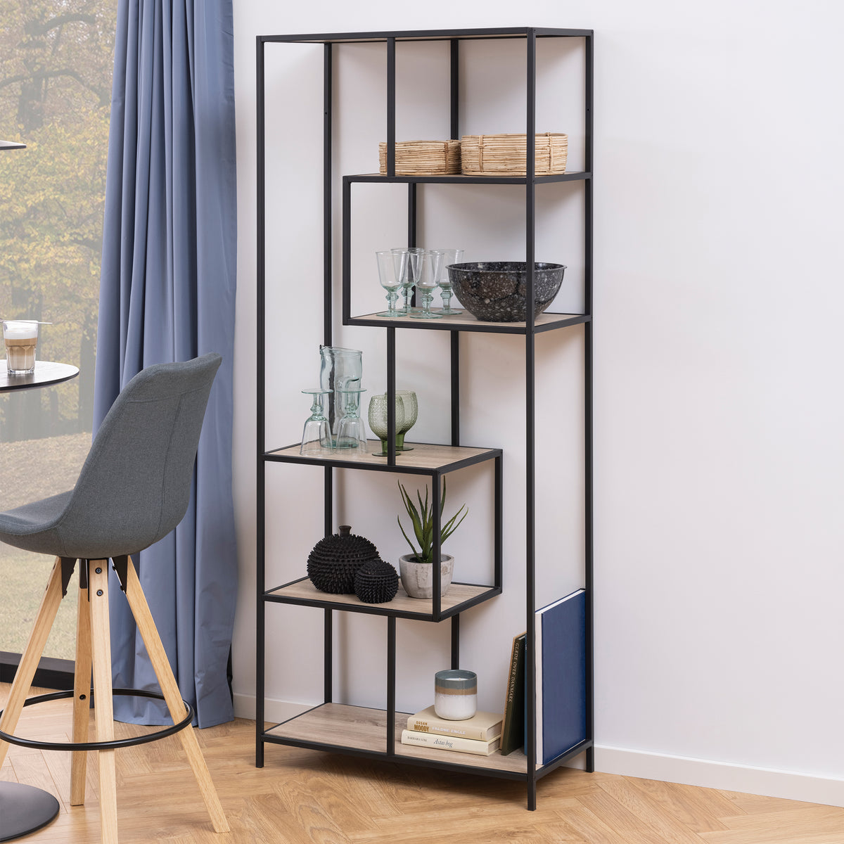 The Skeleto bookcase (77x185), in Sonoma Oak, by the window with a blue curtain, next to a chair – a bookcase for books and glassware in the living room