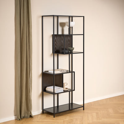 A Skeleto bookcase (77x185), in black melamine, against a light-coloured wall next to a linen curtain, with ceramics and books on the shelves