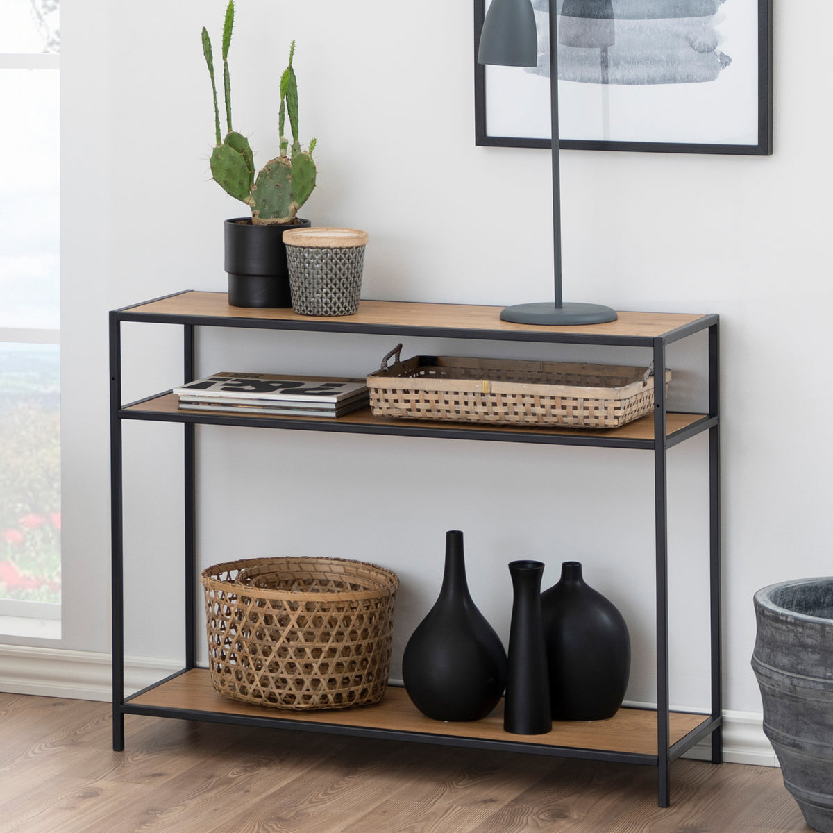 A Skeleto 100x79 Wild Oak console table against the wall, with a cactus, a lamp, baskets and black vases on the bottom shelf